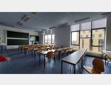 Klassenzimmer mit mehreren rechteckigen Tischen und Holzstühlen, einer großen Kreidetafel, einem Flachbildschirm sowie großen, bodentiefen Fenstern, durch die ein benachbartes Gebäude sichtbar ist. Die Raumbeleuchtung besteht aus Deckenleuchten, und der Boden ist dunkelblau.