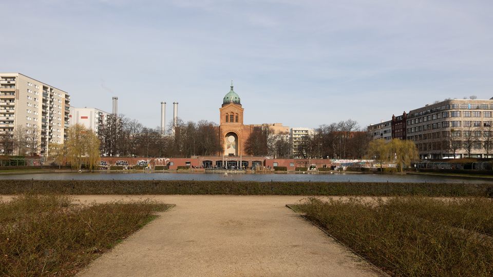 Blick über einen städtischen Parkteich mit einem geraden Weg im Vordergrund; gegenüber steht ein markanter Backsteinturm mit grüner Kuppel und großer Rundöffnung. Links und rechts rahmen mehrgeschossige Wohn- und Bürogebäude sowie kahle Bäume das Ufer.