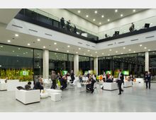 Großer, moderner Innenraum eines Veranstaltungsortes mit einer hohen Decke, Glasfassaden und einer Galerieebene. Der Raum ist mit weißen Sofas, Tischen und Stühlen eingerichtet, an denen Personen in Gespräche vertieft sind; im Hintergrund befinden sich Pflanzen und digitale Bildschirme.