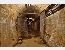 Ein schmaler, gewölbter Tunnel aus gelblich verfärbten Backsteinen mit technischer Infrastruktur, darunter blaue Rohre und Gittertüren. Der Boden ist feucht, und eine Beleuchtungslinie zieht sich entlang der Decke.