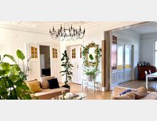 Helles Wohnzimmer mit Holzboden, großen weißen Doppeltüren und einem Kronleuchter. Im Raum stehen Pflanzen, ein beiges Sofa, ein Couchtisch mit Glasplatte sowie ein großer goldgerahmter Spiegel an der Wand.