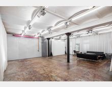 Weitläufiges, industriell gestaltetes Studio mit weiß gestrichener Gewölbedecke, großen Stahlträgern und einem abgenutzten Betonboden. Der Raum ist mit weißen Vorhängen, einer schwarzen Couchgarnitur, einem Spiegel und Beleuchtungseinrichtung ausgestattet.