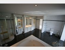 Luxuriöses Badezimmer mit schwarzem glänzendem Boden, einem großen Whirlpool im Vordergrund, einer Glasdusche mit goldenen Verzierungen auf der linken Seite und einem Doppelwaschbecken mit Spiegel und eingebauten Ablagen in der Mitte. Rechts sind eine Toilette und ein Handtuchheizkörper zu sehen, umgeben von weißen Wandfliesen.