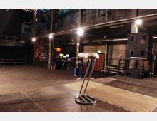 Industriehalle mit einer Bühne, Baustrahlern und einer erhöhten Galerieebene. Im Vordergrund stehen zwei verchromte Barhocker, im Hintergrund sind eine Bar, Lautsprecher und verschiedene metallische Strukturen zu sehen.