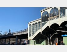 Außenansicht eines U-Bahnhofsgebäudes mit gewölbten Fenstern im historischen Stil, darunter der Eingang zum U-Bahnhof Görlitzer Bahnhof. Eine erhöhte Bahntrasse verläuft links neben dem Gebäude, während Fußgänger und Autos im Straßenbereich darunter zu sehen sind.