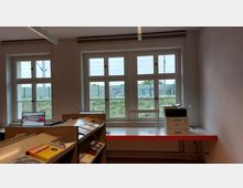 Ein heller Raum mit großen Sprossenfenstern, durch die ein Zaun und eine dahinterliegende Grünfläche mit Graffiti zu sehen sind. Im Vordergrund befinden sich Bücherregale mit Zeitschriften und Büchern sowie ein Schreibtisch mit einem Drucker und einem Monitor.