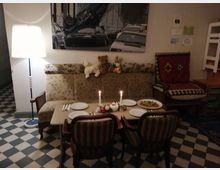 Wohnzimmer mit gemütlicher Einrichtung: Ein gedeckter Esstisch mit vier Tellern, Besteck und Kerzen steht vor einem Sofa mit gemustertem Bezug. Im Hintergrund ist ein großes Wandbild, neben dem eine Stehlampe steht, der Boden ist kariert gefliest.