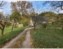 Ländliche Szene mit einem Feldweg, der durch einen Garten mit Obstbäumen führt, und einem Gebäude mit Schindeldach im Hintergrund. Das Gelände ist von grünem Gras und Herbstlaub bedeckt.