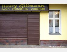 Fassade eines Ladengeschäfts mit geschlossenen braunen Rollläden, gelber Wandfarbe und einem Fenster mit weißer Gardine. Über den Rollläden befindet sich ein Schild mit der Aufschrift "Henry Gillmann Radio · Fernsehen · Zubehör".