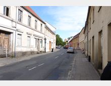 Straßenansicht einer kleinen Altstadt mit schmaler Asphaltstraße, flankiert von leicht verfallenen, mehrstöckigen Gebäuden mit verputzten Fassaden auf der linken Seite und einfachen, gelblichen Häusern auf der rechten Seite. Im Hintergrund sind Bäume und der Turm einer Kirche sichtbar.