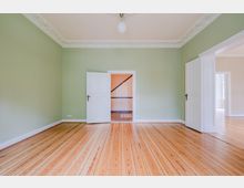 Leerer Wohnraum mit hellgrünen Wänden, breiten Holzdielen und dekorativer Stuckleiste an der Decke mit einfacher Deckenlampe. Zwei weiße Flügeltüren öffnen in einen Flur mit Treppe sowie in einen angrenzenden, hellen Raum.