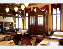 Innenraum eines traditionellen Restaurants mit Holzausstattung; große Fenster mit roten Vorhängen, Tische mit weißen Tischdecken und gepolsterte Stühle. Im Vordergrund sind Deckenleuchten und dekorative, verzierte Holztüren sichtbar.