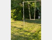 Eine einfache Schaukel mit Metallrahmen steht auf einem Rasen in einem Garten, umgeben von dichten, grünen Sträuchern und Bäumen. Die Szene ist von natürlichem Licht erhellt.