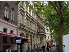 Straßenszene in einem Wohngebiet mit Altbauten und verzierten Fassaden. Links befinden sich Geschäfte mit Markisen, während Fahrräder entlang des Gehwegs abgestellt sind; im Hintergrund stehen dicht belaubte Bäume.