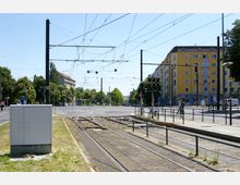 Straßenbahnkreuzung mit Schienen und Oberleitungen in einer urbanen Umgebung. Im Hintergrund sind ein gelbes Wohngebäude, Fußgänger, Bäume und Verkehrssignale zu sehen.