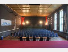 Innenansicht eines Veranstaltungsraums mit Stuhlreihen, die auf eine Bühne ausgerichtet sind. Der Raum hat schwarze Wände, helle Fenster auf der rechten Seite und eine abgehängte Decke; im Hintergrund hängen rote Stoffdekorationen neben Scheinwerfern.