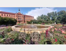 Großer Garten mit einem Springbrunnen im Vordergrund, umgeben von bunten Blumenbeeten. Im Hintergrund steht ein historisches Backsteingebäude mit einer Turmuhr und verzierten Dachgiebeln.