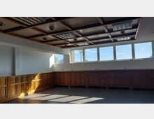 Leerer Raum mit Holzwandvertäfelung und Kassetten-Decke, ausgestattet mit eingebauten Deckenleuchten und Lüftungsgittern. Große Fenster auf der rechten Seite lassen Tageslicht herein und geben den Blick auf den Himmel frei.