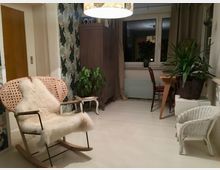 Gemütlicher Wohnraum mit einem Schaukelstuhl aus Holz mit geflochtener Sitzfläche und einem Fell, einem kleinen Holztisch mit Stuhl, sowie mehreren großen Zimmerpflanzen. Im Hintergrund befindet sich ein Fenster mit Vorhängen, ein Schrank und eine helle Wand mit floralem Muster.