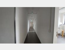 Ein schmaler, weiß gestrichener Flur mit dunklem Boden und mehreren Türen an beiden Seiten. Im Hintergrund ist eine geschlossene Tür mit einem grünen Notausgangsschild darüber sichtbar, rechts befindet sich ein teilweise einsehbares Zimmer mit einem Stuhl und einem Tisch.