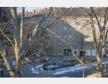 Straßenszene mit einem großen Wohngebäude im Hintergrund, dessen Fassade von mehreren Fenstern durchbrochen ist. Im Vordergrund sind kahle Bäume und ein kleiner Platz mit Schneeresten sowie parkende Autos zu sehen.