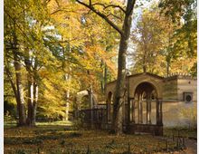 Historische Steinarchitektur mit Bögen und verzierten Details inmitten eines Parks mit herbstlich gefärbtem Laub. Hohe Bäume umgeben die Struktur, deren Umgebung von Laub bedeckt ist.
