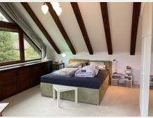 Schlafzimmer in einem Dachgeschoss mit schrägen weißen Wänden und freiliegenden Holzbalken. Ein großes Bett mit heller Bettwäsche steht in der Mitte, daneben Nachttische mit Lampen; links befindet sich ein großes Fenster mit Vorhängen und einer Holzkommode darunter.