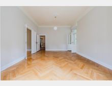 Leerer, heller Wohnraum mit weiß gestrichenen Wänden, Stuckdecke und mittiger Hängeleuchte sowie Fischgrätparkettboden. Mehrere weiße Flügeltüren öffnen zu angrenzenden Räumen und zu einer Fenster-/Balkontür auf der rechten Seite.