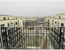 Mehrstöckige, moderne Wohngebäude mit hellgelber Fassade und Balkonen, umgeben von einer zentralen Grünfläche. Der Blick erfolgt von einem Balkon mit schwarzem Geländer; im Hintergrund sind kahle Bäume unter grauem Himmel zu sehen.