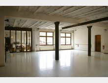 Ein heller Tanz- oder Übungsraum mit weißem Boden, freiliegenden Balken an der Decke und Säulen in der Mitte. Große Fenster mit Holzeinfassung lassen Tageslicht herein, auf der linken Seite befindet sich eine verglaste Wand, die zu einem angrenzenden Raum mit Tischen und Stühlen führt.