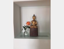 Eine goldene Buddha-Statue sitzt auf einem Sockel in einer weißen Nische, daneben steht eine einzelne orangefarbene Rose in einer kleinen Glasvase.