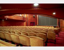 Innenansicht eines Theatersaals mit mehreren Reihen gepolsterter, beige-farbener Sitze, rotem Teppichboden, hölzernen Wänden und Blick auf eine geschlossene Bühne mit schwarzem Vorhang. Oben ist ein darüberliegender Balkon sichtbar.