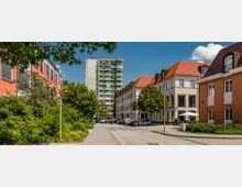 Eine städtische Straße mit modernen Wohn- und Geschäftsgebäuden, darunter ein mehrstöckiges Wohnhaus mit grünen Balkonen im Hintergrund und Häuser mit roten Ziegeldächern im Vordergrund. Am Straßenrand stehen geparkte Autos, umgeben von Bäumen und Grünflächen.