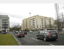 Mehrspurige Straße in einer Stadt mit vorbeifahrenden Autos, flankiert von mehrstöckigen Wohnhäusern mit Balkonen und Gewerbeflächen im Erdgeschoss. Im Hintergrund sind Straßenlaternen, Bäume und Fußgängerampeln zu sehen.