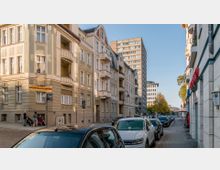 Straßenszene in einer Stadt mit mehrstöckigen Wohngebäuden im Altbau-Stil auf der linken Seite und einem modernen Hochhaus im Hintergrund. Geparkte Autos säumen die Straße, während Passanten mit Kinderwagen auf dem Gehweg gehen.