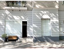 Fassade eines hellgrauen Gebäudes mit zwei großen Fenstern mit geschlossenen Jalousien und einer Glastür in der Mitte. Vor der Tür führen Treppenstufen mit einem Geländer nach oben, daneben steht eine Holzbank und ein Blumenkübel.