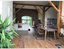 Großer, offener Wohnraum mit sichtbaren Holzbalken und heller Holzdecke. Im Vordergrund ein Esstisch mit Stühlen, im Hintergrund eine offene Küche mit weißen Schränken und ein großes Fenster mit Blick ins Grüne; links und rechts Bücherregale und Zimmervorhänge.