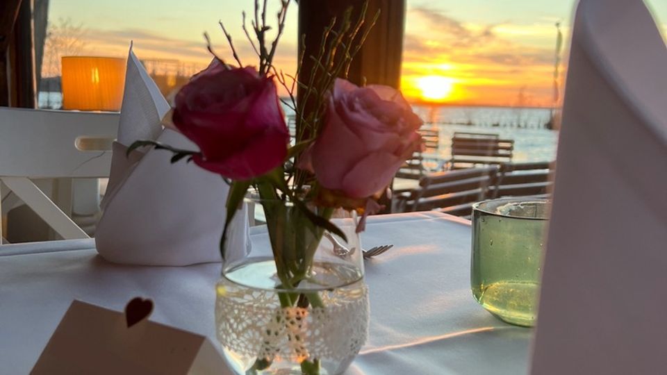 Ein festlich gedeckter Tisch in einem Restaurant mit Blick auf einen Sonnenuntergang über einem Gewässer. Im Vordergrund stehen ein Glas mit rosa Blumen, eine Tischkarte und eine grüne Glasvase, während durch große Holzfenster die Außenlandschaft mit Holzstegen und Spiegelungen der Sonne sichtbar ist.