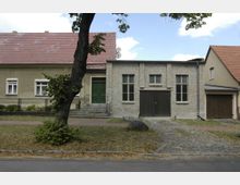 Zwei aneinandergrenzende Gebäude in einer ruhigen Straße, links ein gelbes Wohnhaus mit grünem Eingang und zwei Fenstern, rechts ein graues Gebäude aus Backstein mit großen Fenstern und einer Holztor-Garage. Im Vordergrund ein Gehweg, eine Rasenfläche und ein Baum.