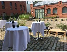 Ein gepflasterter Hof mit rötlichen Backsteingebäuden im Hintergrund. Im Vordergrund stehen runde Stehtische mit weißen Tischdecken, darauf Flaschen Wein und kleine Pflanzen; umgeben von Blumenkübeln und Holzpaletten.