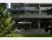 Wohnhaus mit mehreren Balkonen in einem mehrstöckigen Gebäude aus Beton, Glas und Backstein. Vor dem Eingang befinden sich ein gepflasterter Weg, Fahrräder sowie Bäume und Büsche im Vordergrund.