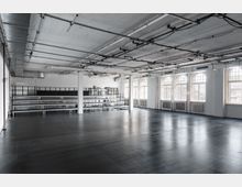 Großer, leerer Loft-Industrieraum mit dunklem Boden, weißen Stützen und sichtbaren Leitungen an der Decke. Entlang einer Seite befinden sich hohe Regalreihen, gegenüber eine Reihe großer Rundbogenfenster mit Heizkörpern darunter.