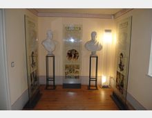 Raum mit zwei Marmorbüsten auf schwarzen Podesten, flankiert von gläsernen Informationstafeln mit Texten und Bildern; Holzboden und dezente Wanddekorationen mit heller Beleuchtung.