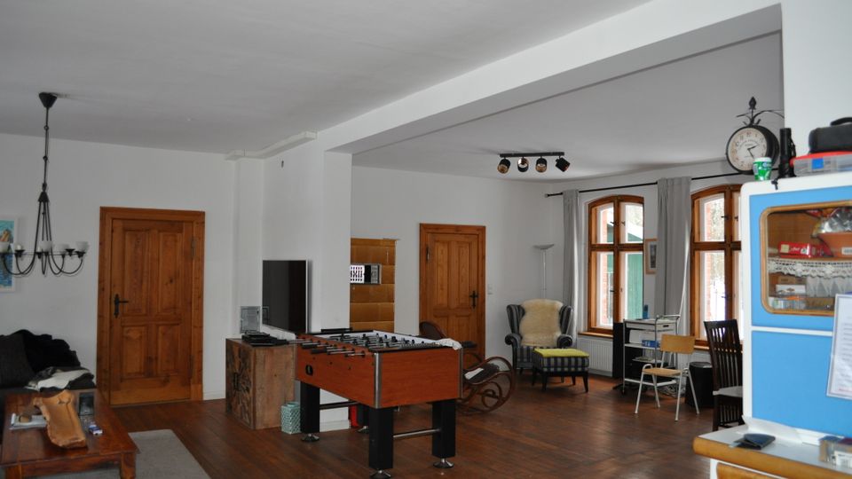 Wohnbereich mit Holzboden, weiß gestrichenen Wänden und mehreren Holztüren. Der Raum ist mit großen Holzrahmenfenstern, einem Tischkicker, einem Sofa, einem Sessel und einem Hängeschrank ausgestattet.