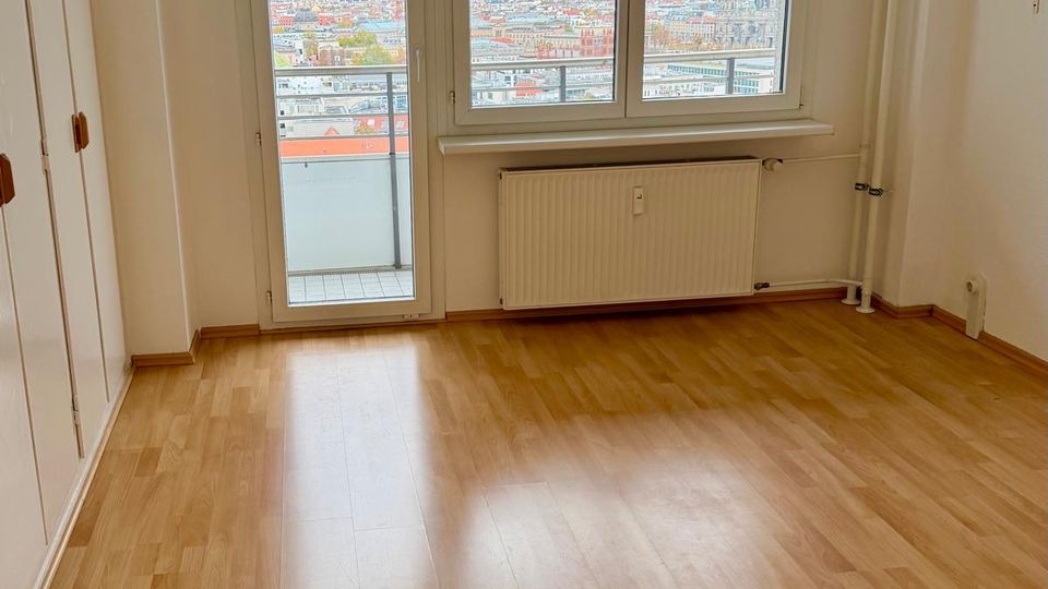 Ein leerer Raum mit hellem Laminatboden, weißen Wänden und einem großen Fenster mit Balkontür, durch das eine Aussicht auf eine Stadtlandschaft unter bewölktem Himmel zu sehen ist. Unter dem Fenster befindet sich ein weißer Heizkörper.