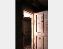 Alte, verwitterte Tür in einem dunklen Raum, auf der das Wort "BAR" mit einem Pfeil handgeschrieben ist. Dahinter ist ein gefliester Raum mit gedämpftem Licht zu sehen.