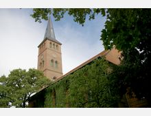 Backsteinkirche mit hohem Turm und spitzem Schieferdach, umgeben von dichtem Laubwerk. Die Kirchenfassade ist teilweise mit Efeu bewachsen, während der Himmel im Hintergrund klar ist.