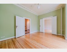 Leerer Altbau-Wohnraum mit grün gestrichenen Wänden, hellem Holzdielenboden und Stuckverzierungen an der Decke. Zwei weiße Doppeltüren führen in angrenzende Räume, rechts sind sichtbare Heizungsrohre an der Wand.