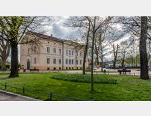 Historisches zweistöckiges Gebäude mit cremefarbener Fassade und zahlreichen Fenstern, umgeben von einem Park mit Rasenflächen, Bäumen und Wegen; im Vordergrund Sitzbänke und Blumenbeete.