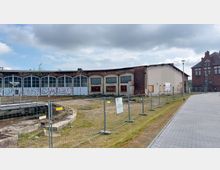 Ehemaliges Industriegebäude mit abgenutzter Fassade und großen, bogenförmigen Fenstern im Vordergrund, teilweise mit Graffiti bedeckt. Der Bereich ist mit einem Bauzaun abgesperrt, und im Hintergrund ist ein historisches rotes Backsteingebäude zu sehen.